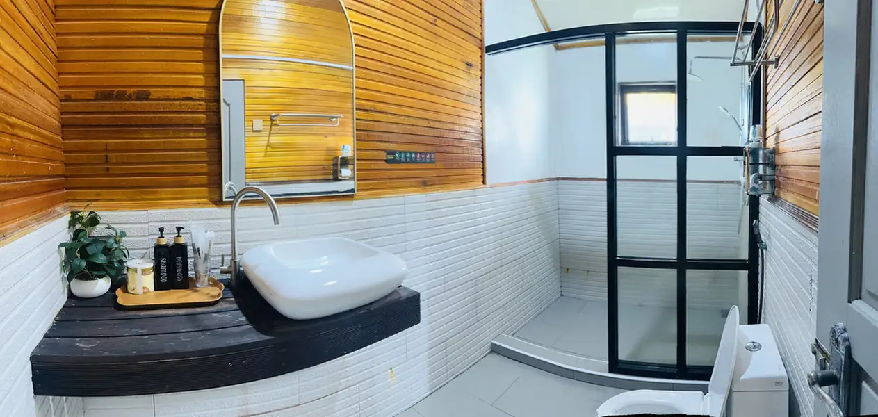 A beautiful bathroom at Dayang Resort detailing warm wood-look wall tiles, a modern vanity with a round mirror, a white ceramic toilet, and a glass-enclosed stall featuring an electric shower.